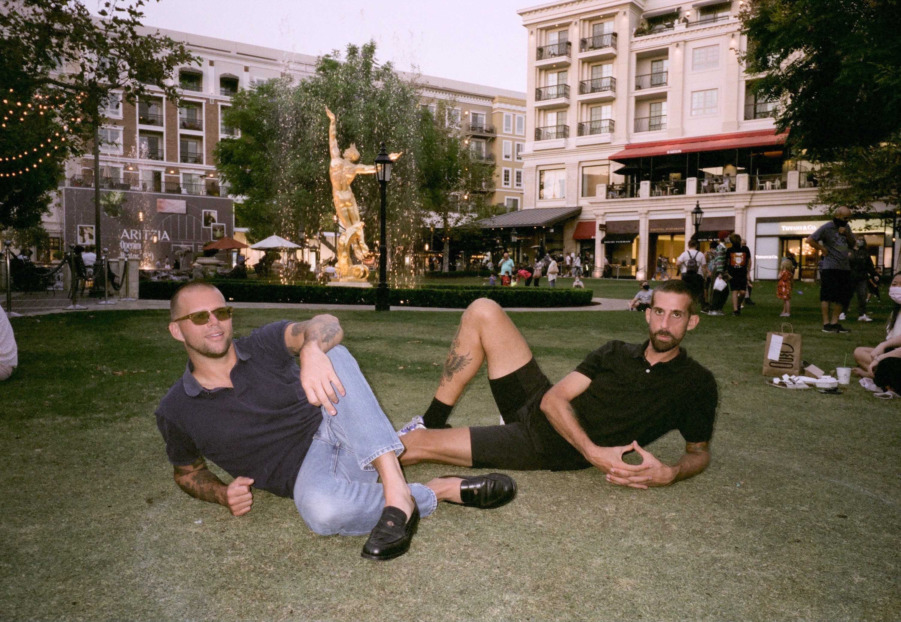 A grainy photograph features How Long Gone hosts Chris Black and Jason Stewart reclining in the grass before a fountain containing a large golden statue of a leaping man.